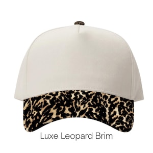 May include: A cream-colored baseball cap featuring a leopard print brim and a small leopard print button at the top. The text Luxe Leopard Brim is printed in black below the cap, which has a structured five-panel design and a curved visor.