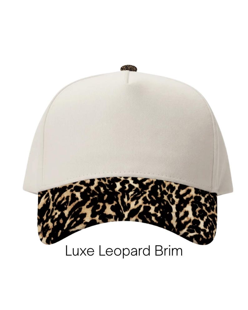 May include: A cream-colored baseball cap featuring a leopard print brim and a small leopard print button at the top. The text Luxe Leopard Brim is printed in black below the cap, which has a structured five-panel design and a curved visor.