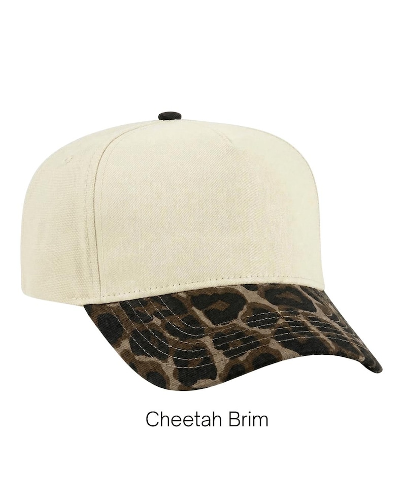 May include: A baseball cap with a cream-colored crown and a black button on top. The brim features a brown and black cheetah print pattern. The text "Cheetah Brim" is printed in black below the cap against a plain white background.