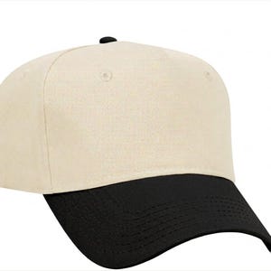 May include: A baseball cap featuring a solid cream-coloured crown and a contrasting black visor. The cap has a structured front panel, a small black button at the top centre, and visible stitching along the curved brim, providing a clean look.