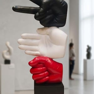 Puede incluir: Una escultura vertical que presenta tres manos apiladas sobre una base rectangular negra. La mano inferior es roja y está cerrada en un puño, la mano central es blanca y está abierta, y la mano superior es negra, formando un signo de paz.