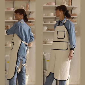 May include: Cream-colored apron with black trim and pockets. The apron is worn over a denim shirt and jeans. The apron has a bib, a large front pocket, and a split at the knees. The apron is tied at the waist with a matching cream-colored tie.