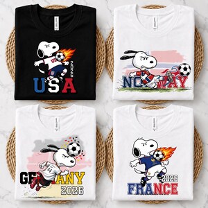 May include: Four t-shirts featuring Snoopy playing soccer. The black shirt says USA 2026, the white shirts feature Norway, Germany 2026, and France 2026. Each shirt displays Snoopy in a different soccer uniform with a soccer ball and country-themed graphics.