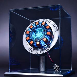 Puede incluir: Una réplica circular de un reactor de arco encerrada en una caja de exhibición de acrílico transparente. El dispositivo presenta un centro azul brillante, bobinas de color cobre y un anillo de metal plateado grabado PROOF THAT TONY STARK HAS A HEART.