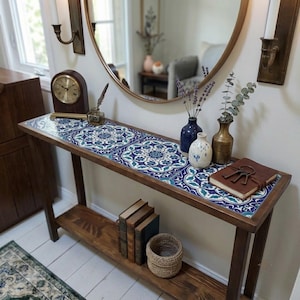 Op de afbeelding: Een rechthoekige houten sidetable met een betegeld blad voorzien van een blauw, wit en groen bloemenmandala-patroon. De tafel heeft een onderste plank met drie boeken en een mand, terwijl op het blad een klok en diverse vazen staan.