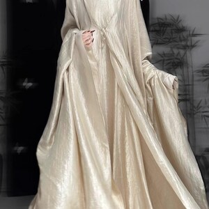 K&ouml;nnte beinhalten: Ein bodenlanges, champagnerfarbenes Kleid mit quadratischem Ausschnitt und einem passenden, lang&auml;rmeligen, vorne offenen &Uuml;berwurf. Der Stoff hat einen dezenten, knittrigen, metallischen Glanz und erzeugt eine flie&szlig;ende, volumin&ouml;se Silhouette mit weichen Falten.