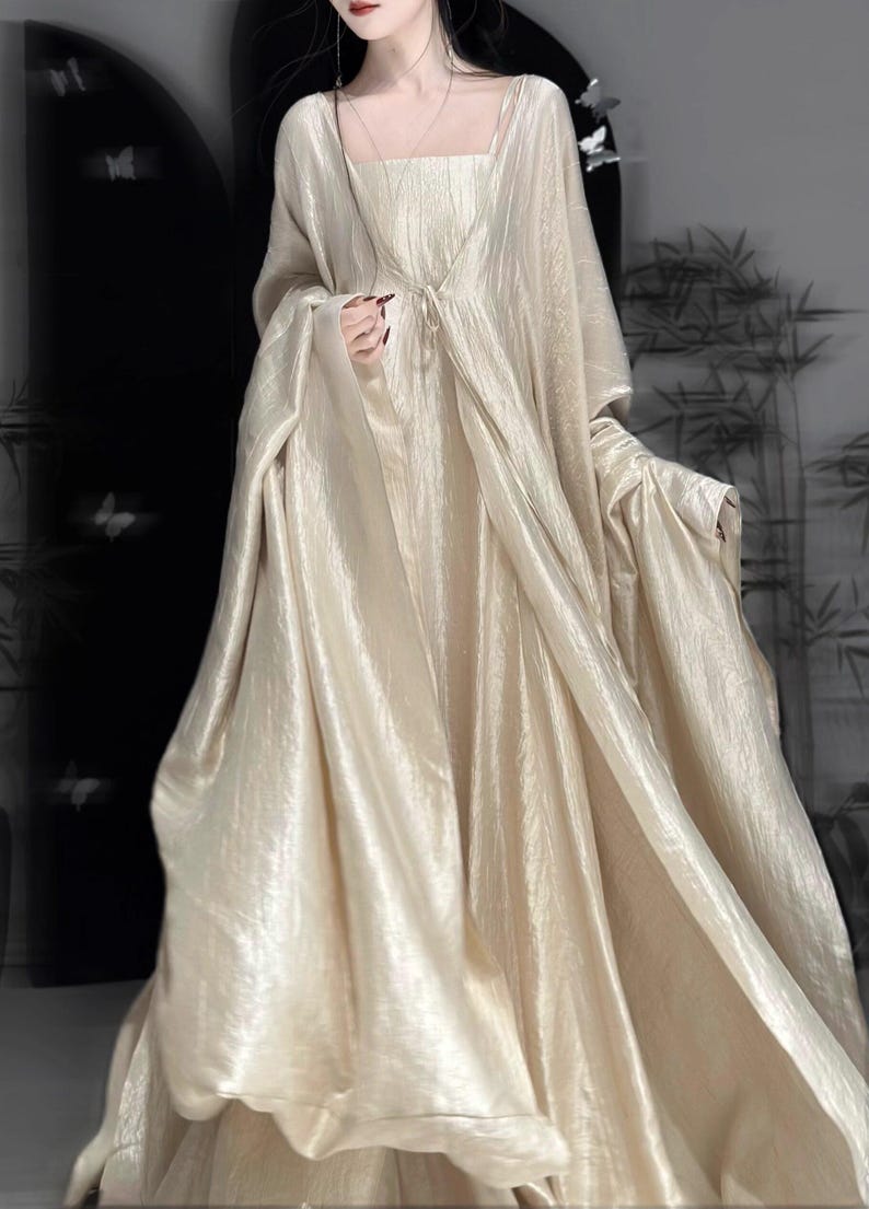 K&ouml;nnte beinhalten: Ein bodenlanges, champagnerfarbenes Kleid mit quadratischem Ausschnitt und einem passenden, lang&auml;rmeligen, vorne offenen &Uuml;berwurf. Der Stoff hat einen dezenten, knittrigen, metallischen Glanz und erzeugt eine flie&szlig;ende, volumin&ouml;se Silhouette mit weichen Falten.