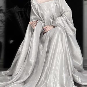 K&ouml;nnte beinhalten: Ein bodenlanges, schimmerndes silbernes Kleid mit quadratischem Ausschnitt und langen, weiten &Auml;rmeln. Der Stoff hat eine zerknitterte, metallische Textur, die locker bis zum Boden f&auml;llt und volumin&ouml;se Falten im gesamten Rock erzeugt.