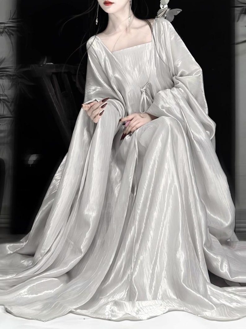 K&ouml;nnte beinhalten: Ein bodenlanges, schimmerndes silbernes Kleid mit quadratischem Ausschnitt und langen, weiten &Auml;rmeln. Der Stoff hat eine zerknitterte, metallische Textur, die locker bis zum Boden f&auml;llt und volumin&ouml;se Falten im gesamten Rock erzeugt.