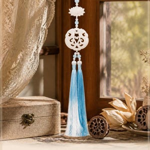 May include: A hanging ornament featuring a white carved lotus flower and a circular openwork jade-like pendant, accented with light blue beads and two long, flowing ombre tassels that transition from white at the top to a vibrant blue at the bottom.