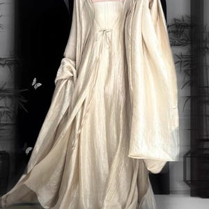 K&ouml;nnte beinhalten: Ein bodenlanges, champagnerfarbenes Kleid mit eckigem Ausschnitt und d&uuml;nnen Spaghettitr&auml;gern. Das Kleidungsst&uuml;ck besteht aus einem strukturierten, schimmernden Stoff mit einer Bindedetail an der Taille und langen, weiten &Auml;rmeln.
