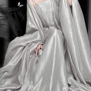 K&ouml;nnte beinhalten: Ein bodenlanges, schimmerndes silbernes Kleid mit quadratischem Ausschnitt und d&uuml;nnen Spaghettitr&auml;gern. Das Kleidungsst&uuml;ck verf&uuml;gt &uuml;ber einen volumin&ouml;sen, flie&szlig;enden Rock und weite, drapierte &Auml;rmel aus leichtem, metallisch texturiertem Stoff.