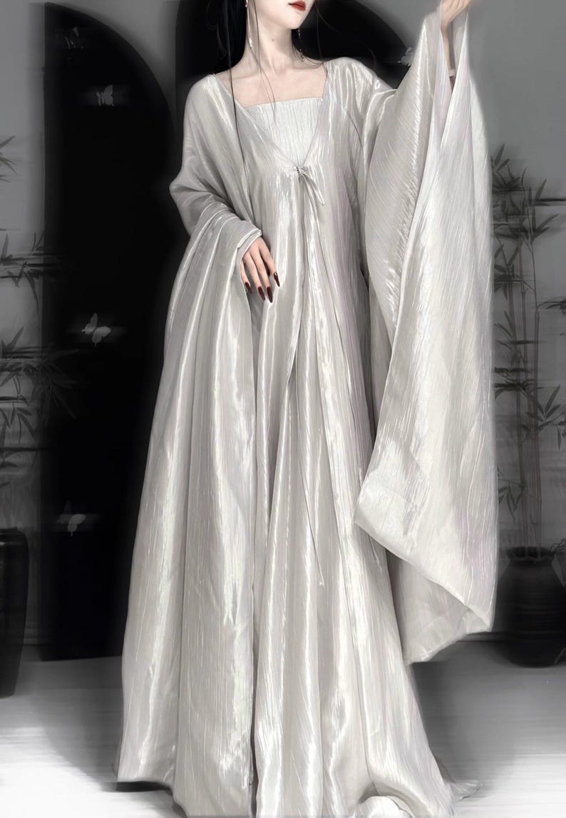 K&ouml;nnte beinhalten: Ein bodenlanges, schimmerndes silbernes Kleid mit quadratischem Ausschnitt, plissiertem Oberteil und langen, weiten, flie&szlig;enden &Auml;rmeln. Das Kleidungsst&uuml;ck besteht aus einem leichten, reflektierenden Stoff, der bis zum Boden f&auml;llt und eine volumin&ouml;se Silhouette erzeugt.