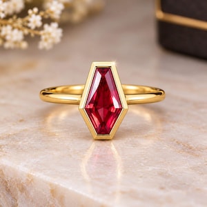 Peut inclure: Une bague en or avec une pierre précieuse rouge en forme de cercueil sertie dans un chaton lisse. L'anneau est fin et poli, mettant en valeur une pierre cramoisie vibrante aux multiples facettes qui captent la lumière avec éclat.