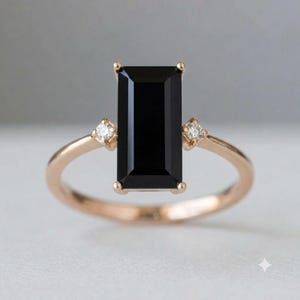 Könnte beinhalten: Ein Ring aus Roségold mit einem großen, rechteckigen, im Smaragdschliff geschliffenen schwarzen Edelstein als Mittelstein. Die Ringschiene ist auf jeder Seite mit einem kleinen, runden, klaren weißen Diamanten in Vier-Krappen-Fassung besetzt.