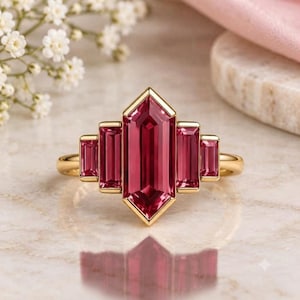 Könnte beinhalten: Ein Goldring mit einem großen, zentralen, vertikalen, sechseckig geschliffenen, tiefrosa Edelstein, der auf jeder Seite von zwei kleineren, im Baguetteschliff geschliffenen rosa Edelsteinen flankiert wird, gefasst in ein poliertes Goldband.