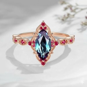 Può includere: Un anello in oro rosa con una gemma centrale taglio marquise che cambia colore in tonalità blu e viola. La montatura è decorata con piccole pietre rotonde di colore rosso vivace disposte in una cornice metallica dal design filigranato.
