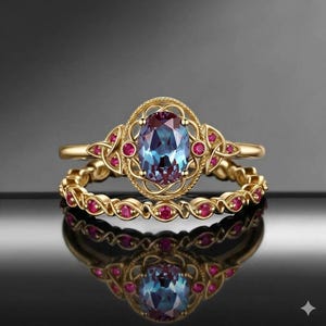 Può includere: Un set di anelli in oro con una pietra preziosa ovale che cambia colore al centro, circondata da un intricato lavoro in filigrana e piccole pietre rosse rotonde. La fascia è decorata con un motivo continuo di piccole pietre rosse incastonate.