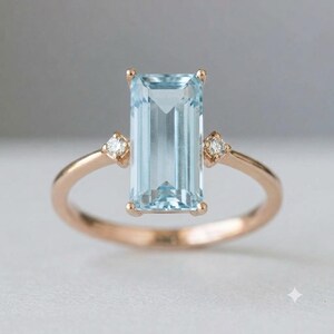 Könnte beinhalten: Ein Ring aus Roségold mit einem großen, rechteckigen, hellblauen Edelstein im Smaragdschliff als Mittelstein, flankiert von zwei kleinen, runden, klaren Akzentsteinen auf beiden Seiten der Ringschiene in einer polierten Fassung.