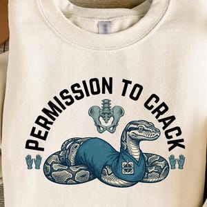 May include: A cream-coloured crewneck sweatshirt featuring a graphic of a snake wearing a blue scrub top with a chiropractor badge. Above the snake is a human pelvis illustration and the text PERMISSION TO CRACK, flanked by two blue glove icons.