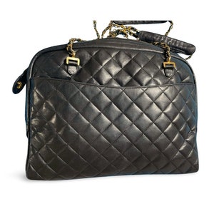 May include: Black quilted leather travel bag with gold-tone chain straps and hardware. The bag features a diamond pattern and a top handle. The bag has a rectangular shape and a zip closure.