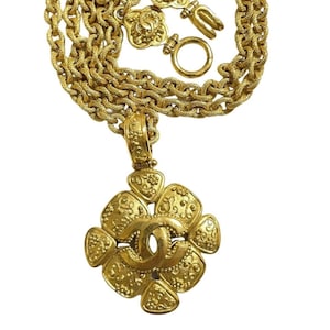 May include: A gold-tone chain necklace with a large, ornate pendant featuring the Chanel logo. The pendant is shaped like a flower with intricate details and a textured finish.