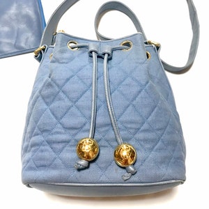 Può includere: Una borsa a secchiello trapuntata blu chiaro con chiusura a coulisse e tracolla a catena dorata. La borsa ha due accenti circolari dorati sul davanti.