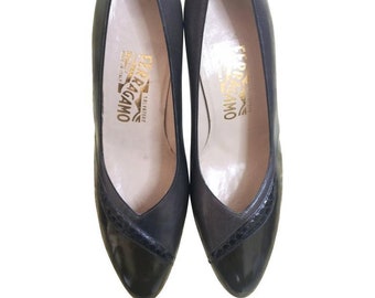 May include: A pair of black leather pumps with a pointed toe and a small heel. The shoes have a black and white trim around the toe and a gold logo on the insole that reads "Salvatore Ferragamo Firenze Made in Italy".