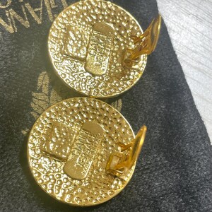 May include: A pair of gold-toned clip-on earrings with a circular design. The earrings feature a textured surface and the word "VERSACE" is visible on the back of each earring.