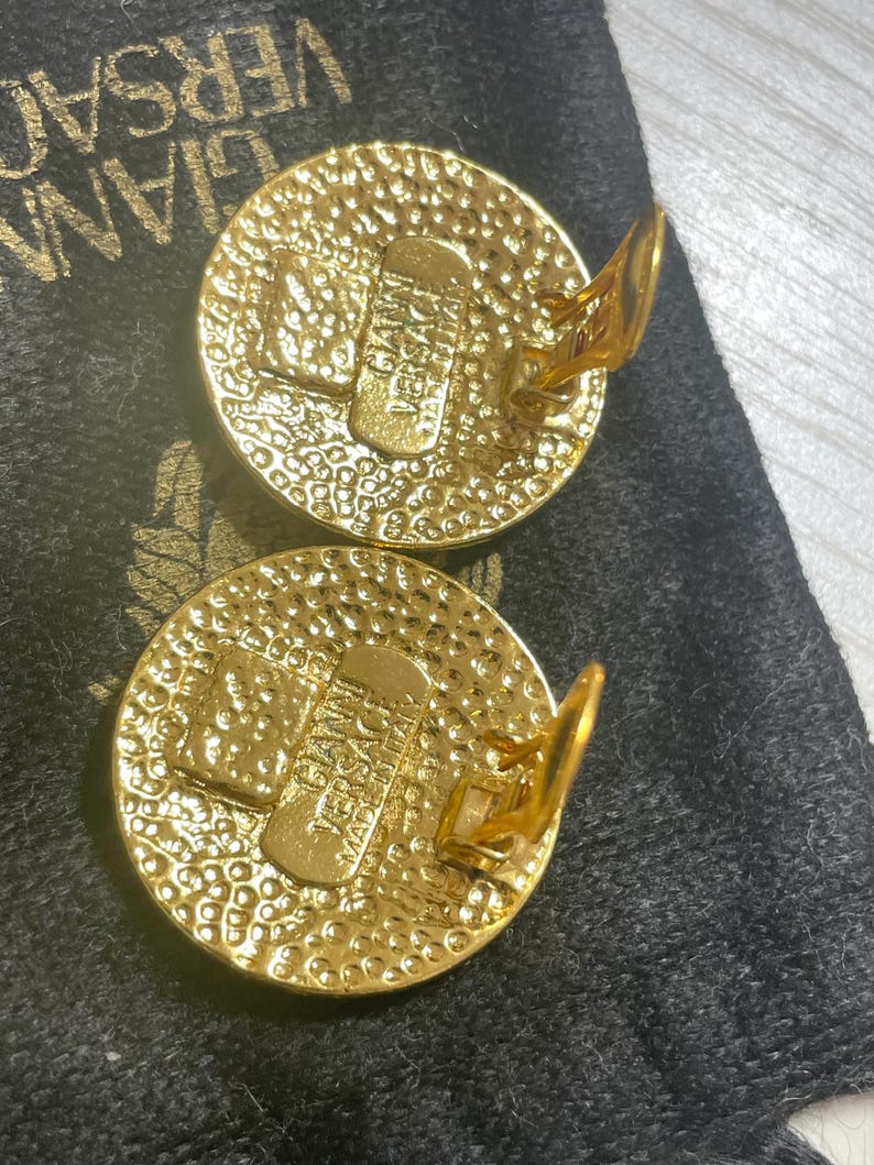 May include: A pair of gold-toned clip-on earrings with a circular design. The earrings feature a textured surface and the word "VERSACE" is visible on the back of each earring.