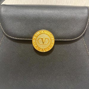 Puede incluir: Bolso de mano negro con un emblema circular dorado con el nombre de la marca "Valentino" y un logotipo "V". El bolso tiene un cierre de solapa y un asa superior. Costuras visibles en color claro.