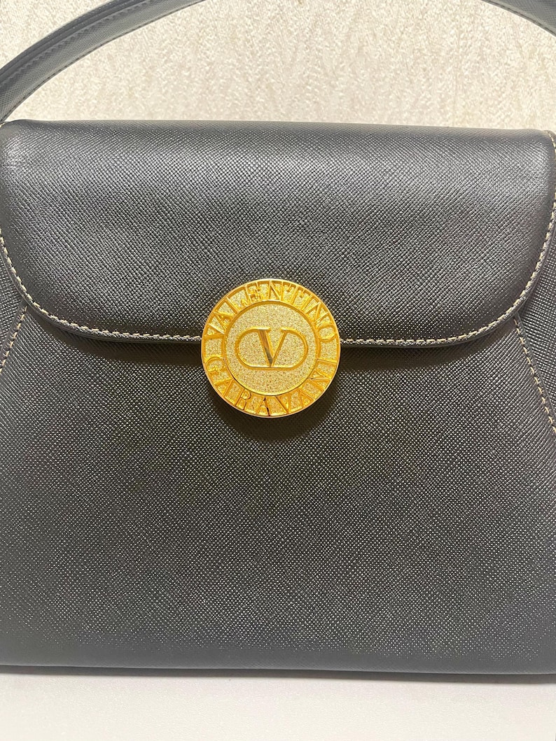 Puede incluir: Bolso de mano negro con un emblema circular dorado con el nombre de la marca "Valentino" y un logotipo "V". El bolso tiene un cierre de solapa y un asa superior. Costuras visibles en color claro.