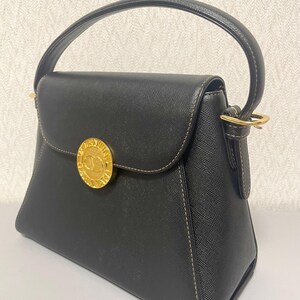 Puede incluir: Bolso de mano negro con asa superior y cierre dorado. El bolso presenta un emblema circular dorado en la parte delantera y costuras en contraste. El bolso tiene una forma estructurada y un cierre de solapa.