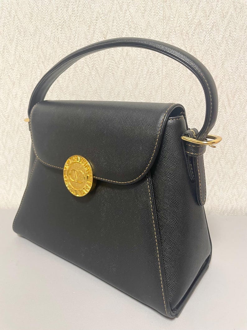 Puede incluir: Bolso de mano negro con asa superior y cierre dorado. El bolso presenta un emblema circular dorado en la parte delantera y costuras en contraste. El bolso tiene una forma estructurada y un cierre de solapa.