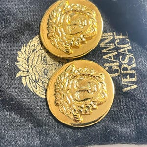 May include: Two gold-toned metal buttons with a raised design of a face with a crown of leaves. The buttons are on a black velvet pouch with the text "Gianni Versace" printed on it.