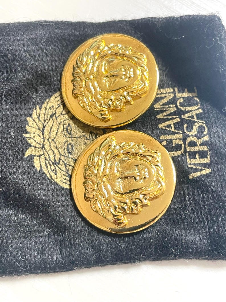 May include: Two gold-toned metal buttons with a raised design of a face with a crown of leaves. The buttons are on a black velvet pouch with the text "Gianni Versace" printed on it.