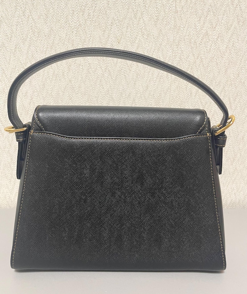 Puede incluir: Bolso de mano negro con asa curva y herrajes dorados. El bolso tiene forma rectangular con cierre de solapa y costuras visibles. El asa est&aacute; sujeta al bolso con aros dorados.