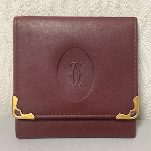 May include: A burgundy leather wallet with gold-toned corner accents. The wallet features an embossed logo in the center and a fold-over closure. The wallet is a compact size.