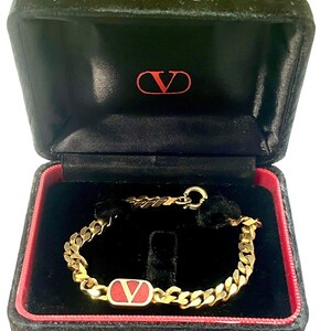 K&ouml;nnte beinhalten: Ein goldfarbenes Kettenarmband mit rotem und goldenem Akzent, pr&auml;sentiert in einer schwarzen Samtbox mit rotem Rand. Die Box hat ein rotes Logo auf der Innenseite des Deckels. Es ist ein luxuri&ouml;ses Schmuckst&uuml;ck.