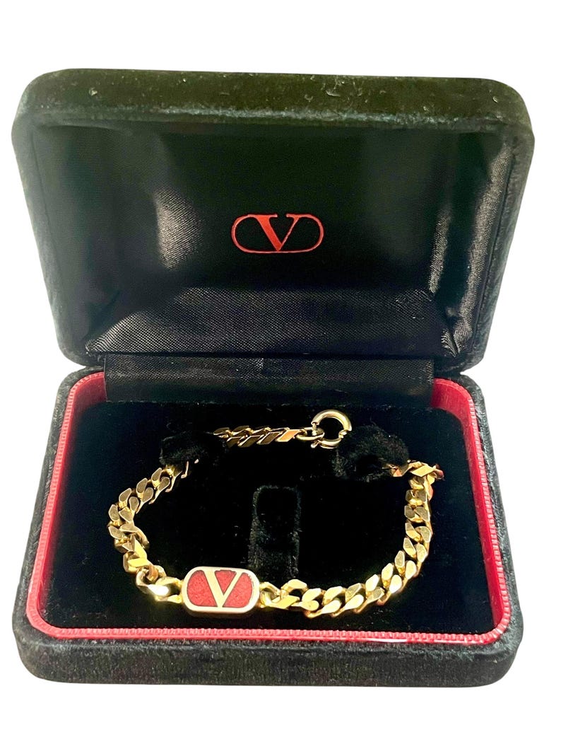 K&ouml;nnte beinhalten: Ein goldfarbenes Kettenarmband mit rotem und goldenem Akzent, pr&auml;sentiert in einer schwarzen Samtbox mit rotem Rand. Die Box hat ein rotes Logo auf der Innenseite des Deckels. Es ist ein luxuri&ouml;ses Schmuckst&uuml;ck.