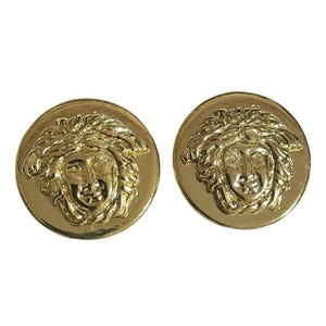 May include: A pair of gold-tone earrings featuring a raised, detailed design of a Medusa head.