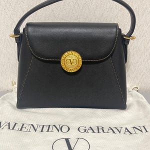 Puede incluir: Bolso de mano negro con un emblema circular dorado con un logotipo "V". El bolso tiene un asa superior y un cierre de solapa. El bolso est&aacute; sobre una bolsa de tela blanca con el texto "VALENTINO GARAVANI" y "LE BORSE".