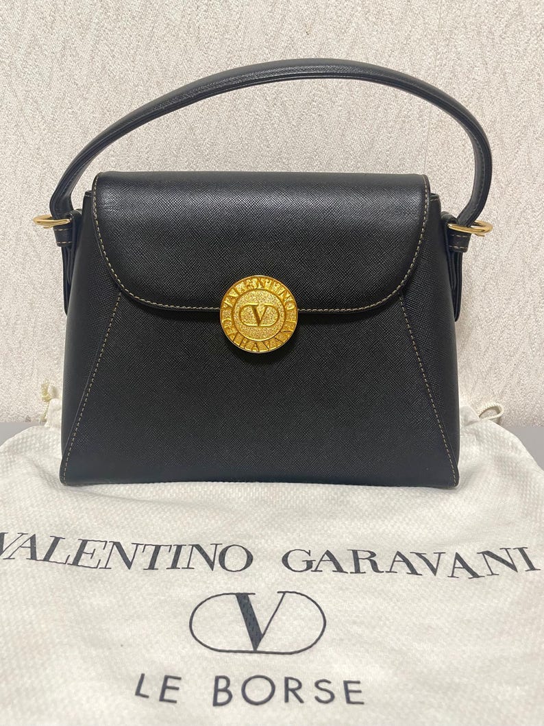 Puede incluir: Bolso de mano negro con un emblema circular dorado con un logotipo "V". El bolso tiene un asa superior y un cierre de solapa. El bolso est&aacute; sobre una bolsa de tela blanca con el texto "VALENTINO GARAVANI" y "LE BORSE".