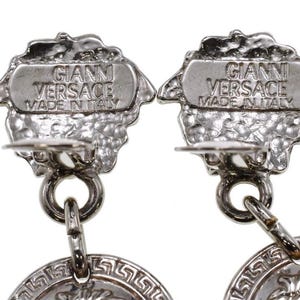 Pode incluir: Close-up de dois brincos Gianni Versace prateados. Cada brinco apresenta um topo texturizado e ornamentado com o nome da marca e o texto "Made in Italy". Os brincos t&ecirc;m um design circular com um padr&atilde;o de chave grega.