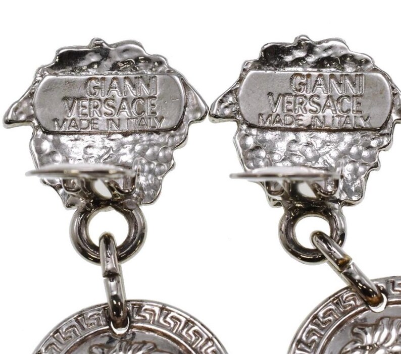 Pode incluir: Close-up de dois brincos Gianni Versace prateados. Cada brinco apresenta um topo texturizado e ornamentado com o nome da marca e o texto "Made in Italy". Os brincos t&ecirc;m um design circular com um padr&atilde;o de chave grega.