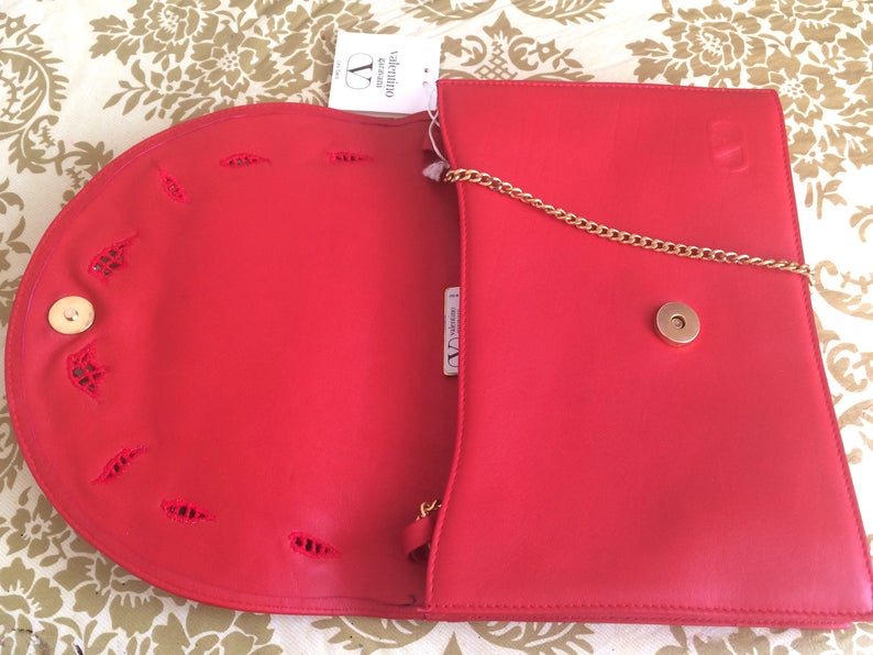 May include: Red leather clutch purse with a gold chain strap and a Valentino Garavani logo tag. The purse has a flap closure and a decorative cut-out design.