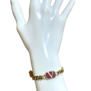K&ouml;nnte beinhalten: Goldfarbenes Kettenarmband mit einem rechteckigen Anh&auml;nger. Der Anh&auml;nger hat einen roten Hintergrund und ein silbernes "V"-Logo. Das Armband hat ein klassisches Panzerketten-Design und einen sicheren Verschluss.