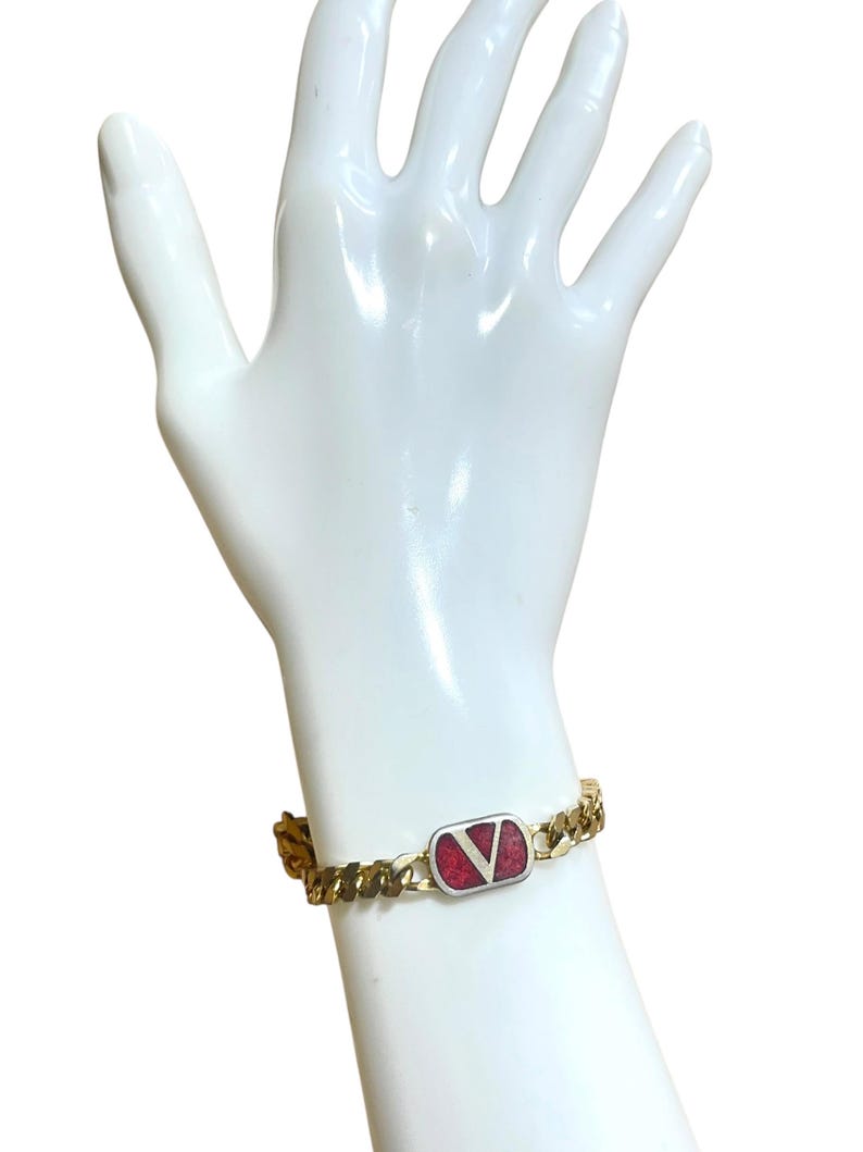 K&ouml;nnte beinhalten: Goldfarbenes Kettenarmband mit einem rechteckigen Anh&auml;nger. Der Anh&auml;nger hat einen roten Hintergrund und ein silbernes "V"-Logo. Das Armband hat ein klassisches Panzerketten-Design und einen sicheren Verschluss.