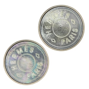 May include: Two circular buttons with a metallic rim. Each button features an iridescent mother-of-pearl center engraved with the text HERMÈS PARIS and two diamond-shaped symbols. The surface of the buttons displays a reflective, pearlescent finish.