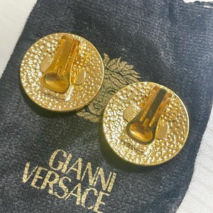 May include: A pair of gold-tone clip-on earrings with a textured circular design. The earrings are in a black velvet pouch with the text "GIANNI VERSACE" printed on it.